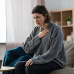 oman sitting on a couch with one hand on her chest, appearing short of breath — representing lingering cardiovascular effects after COVID-19.