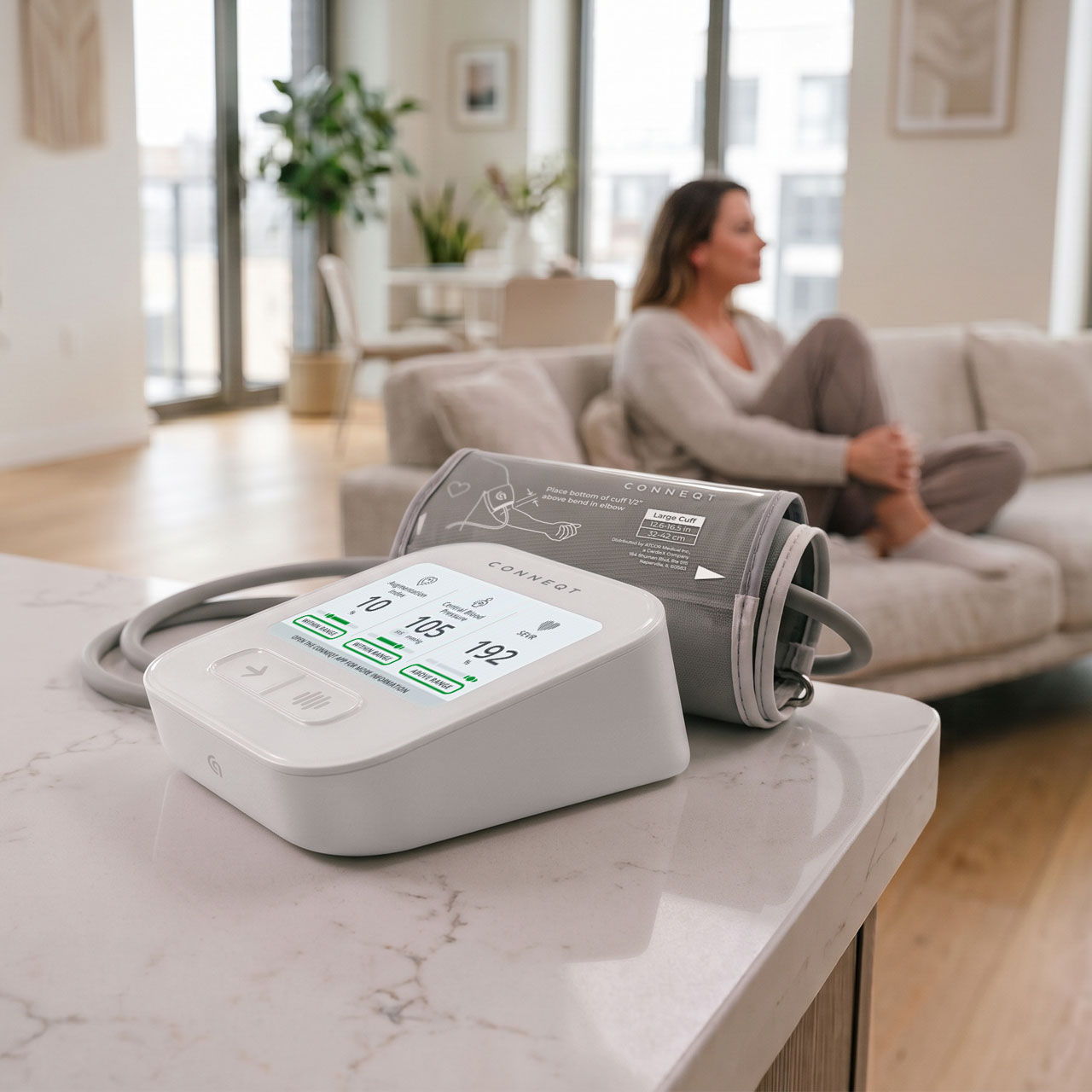 CONNEQT Pulse and its cuff on a table in a bright living room, with a woman relaxing in the background